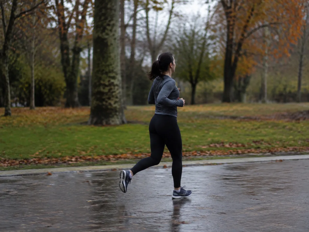 Dorosła kobieta w zwykłym stroju sportowym biegnie spokojnym tempem szeroką ścieżką w miejskim parku o poranku, ujęcie z boku z dystansu