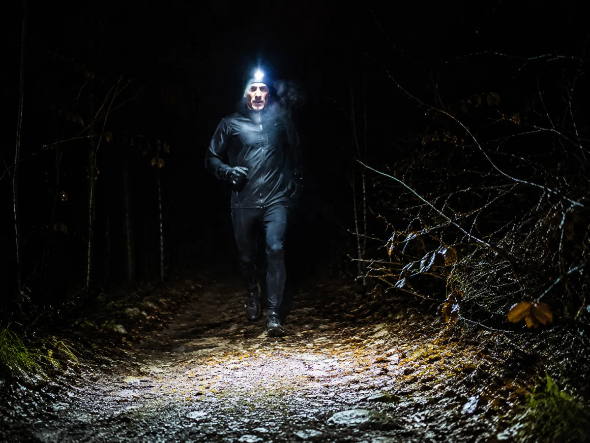 Biegacz w czołówce na nocnej leśnej ścieżce widziany z przodu, szeroka wiązka światła oświetla podłoże kilka metrów przed nim i rozmywa się w ciemności