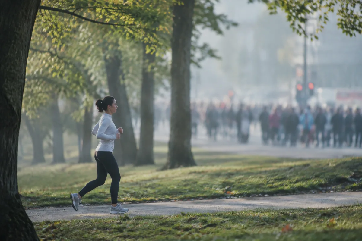 Biegacz truchta spokojnie parkową alejką, w tle za nim nieostry tłum ludzi zebranych przy ulicy, poranne światło