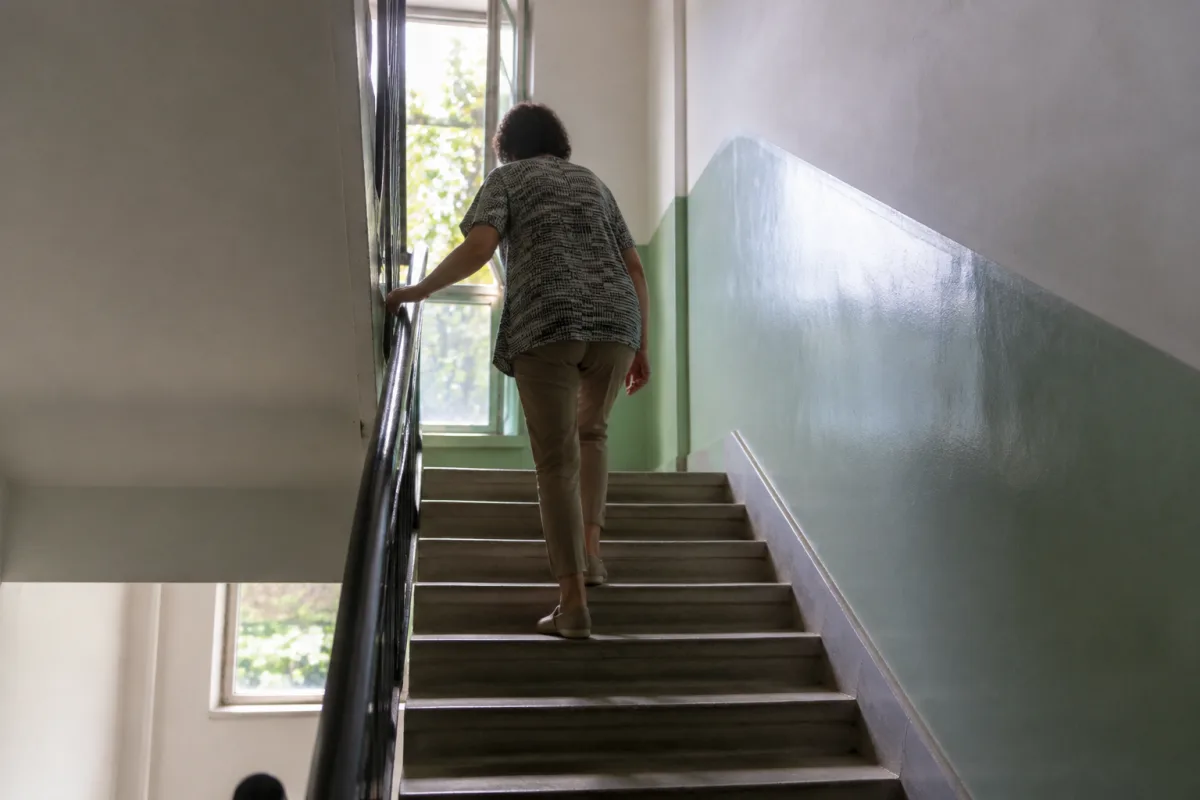 Klatka schodowa w bloku mieszkalnym, osoba w codziennym ubraniu wchodzi po schodach, dłoń na poręczy, światło dzienne z okna na półpiętrze