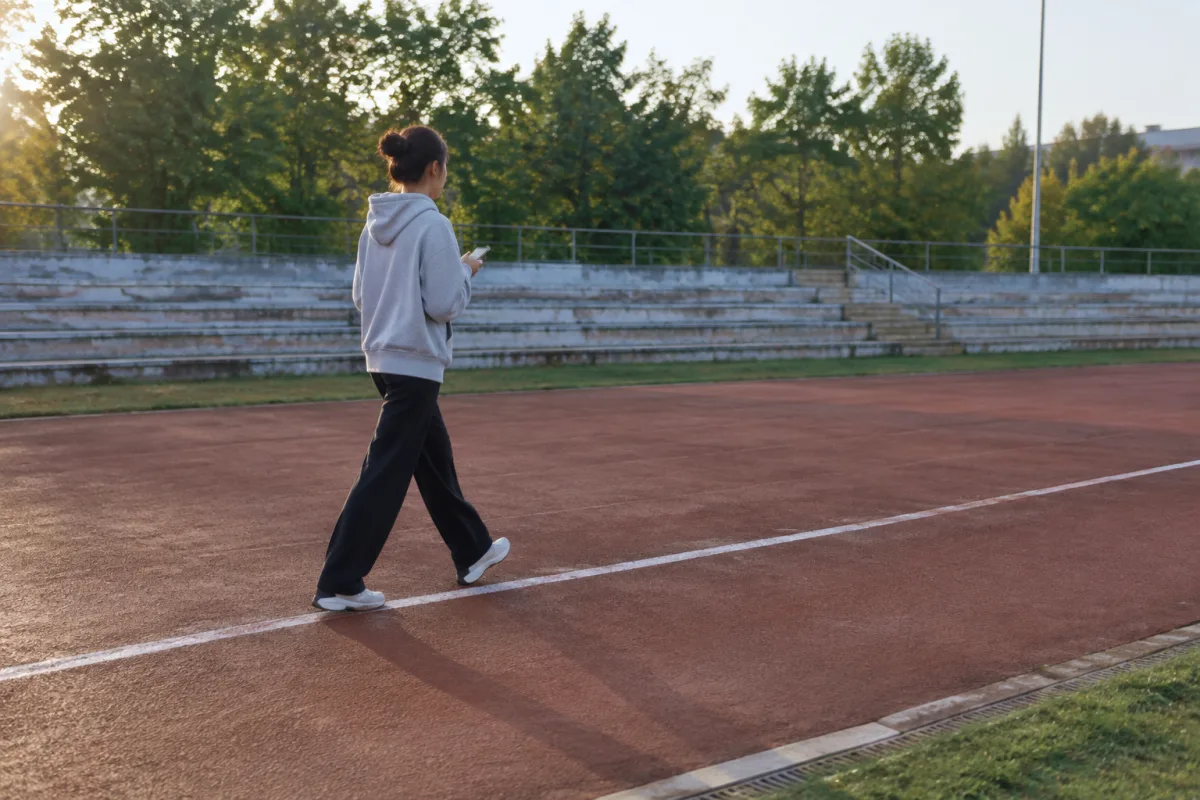 Osoba w codziennym ubraniu idzie wzdłuż białej linii na prostej osiedlowej bieżni stadionu, ujęcie z boku i lekko z tyłu, telefon trzymany w dłoni, poranne światło, puste trybuny w tle
