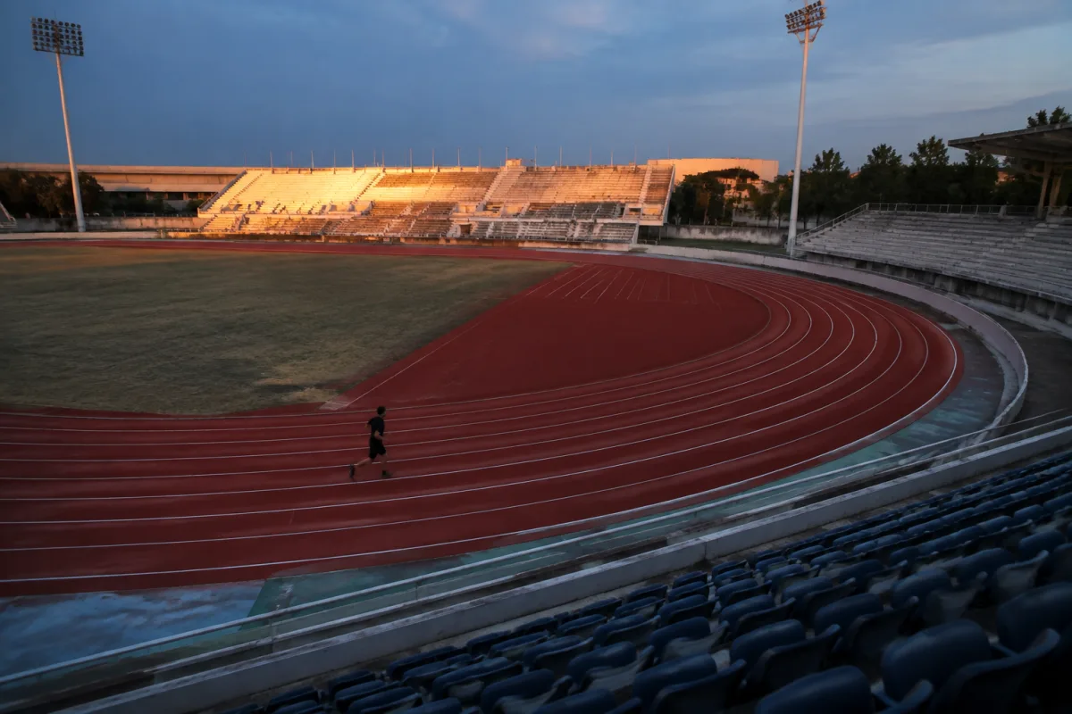 Pusty stadion lekkoatletyczny o zmierzchu, pojedynczy biegacz na dalszym łuku bieżni, kadr obejmuje cały owal z góry