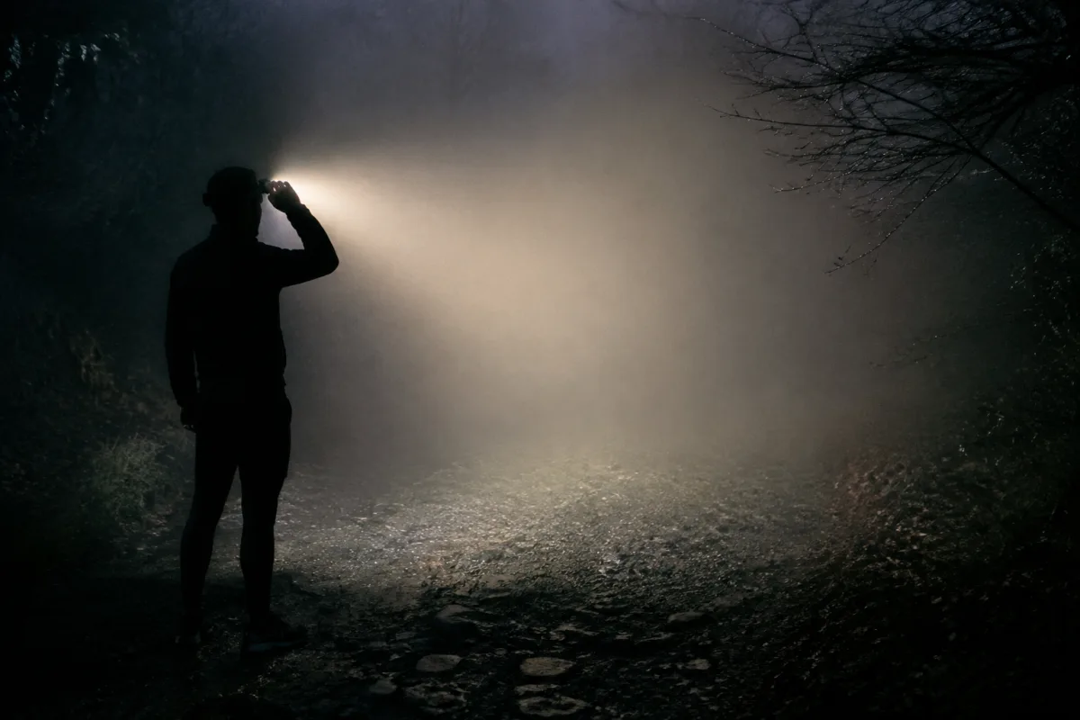 Wiązka czołówki w gęstej mgle w nocnym lesie, światło rozprasza się na kroplach i tworzy jasną białą ścianę zamiast oświetlać ścieżkę