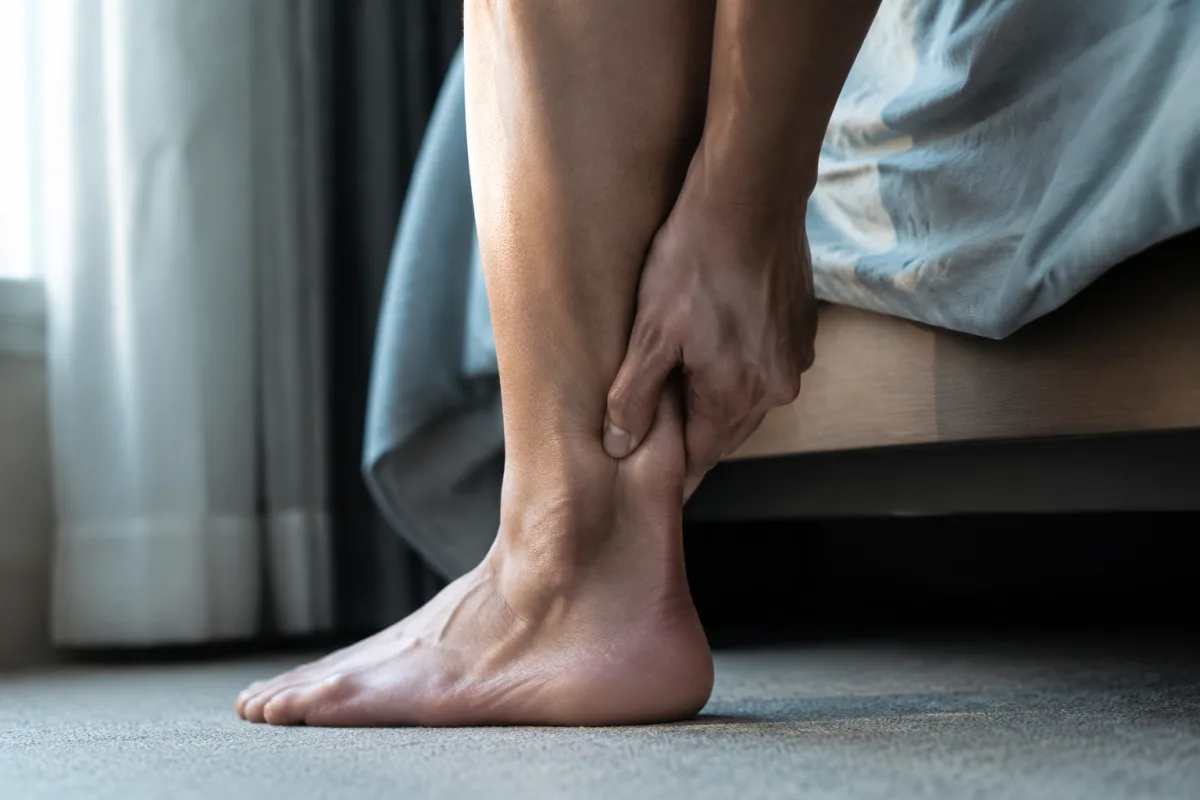 Zbliżenie na łydkę i piętę dorosłej osoby siedzącej na krawędzi łóżka, dłoń uciska ścięgno Achillesa, poranne światło z okna